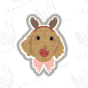 Puede incluir: Un cortador de galletas con forma de cabeza de perro, con cuernos de reno y un lazo rosa. El perro tiene pelaje marrón, nariz roja y ojos negros. El cortador está delineado en gris.