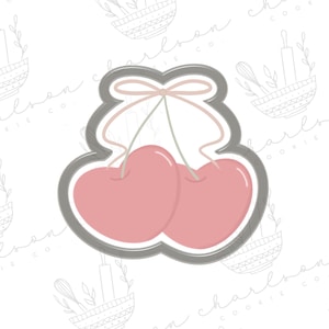 Puede incluir: Un cortador de galletas gris en forma de dos cerezas con tallos y un lazo. Las cerezas son de color rosa.