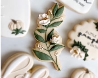 May include: A cookie decorated with a green leafy branch with white flowers and gold accents. The cookie is a light brown color and has a smooth surface. The cookie is a part of a set of cookies with various designs.