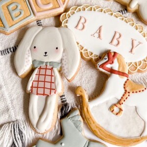 May include: A set of six decorated sugar cookies featuring baby-themed designs. The cookies are decorated with pastel colours and include a rocking horse, a baby rattle, a bunny, a onesie, and blocks that spell "ABC". One cookie has a banner that says "BABY".