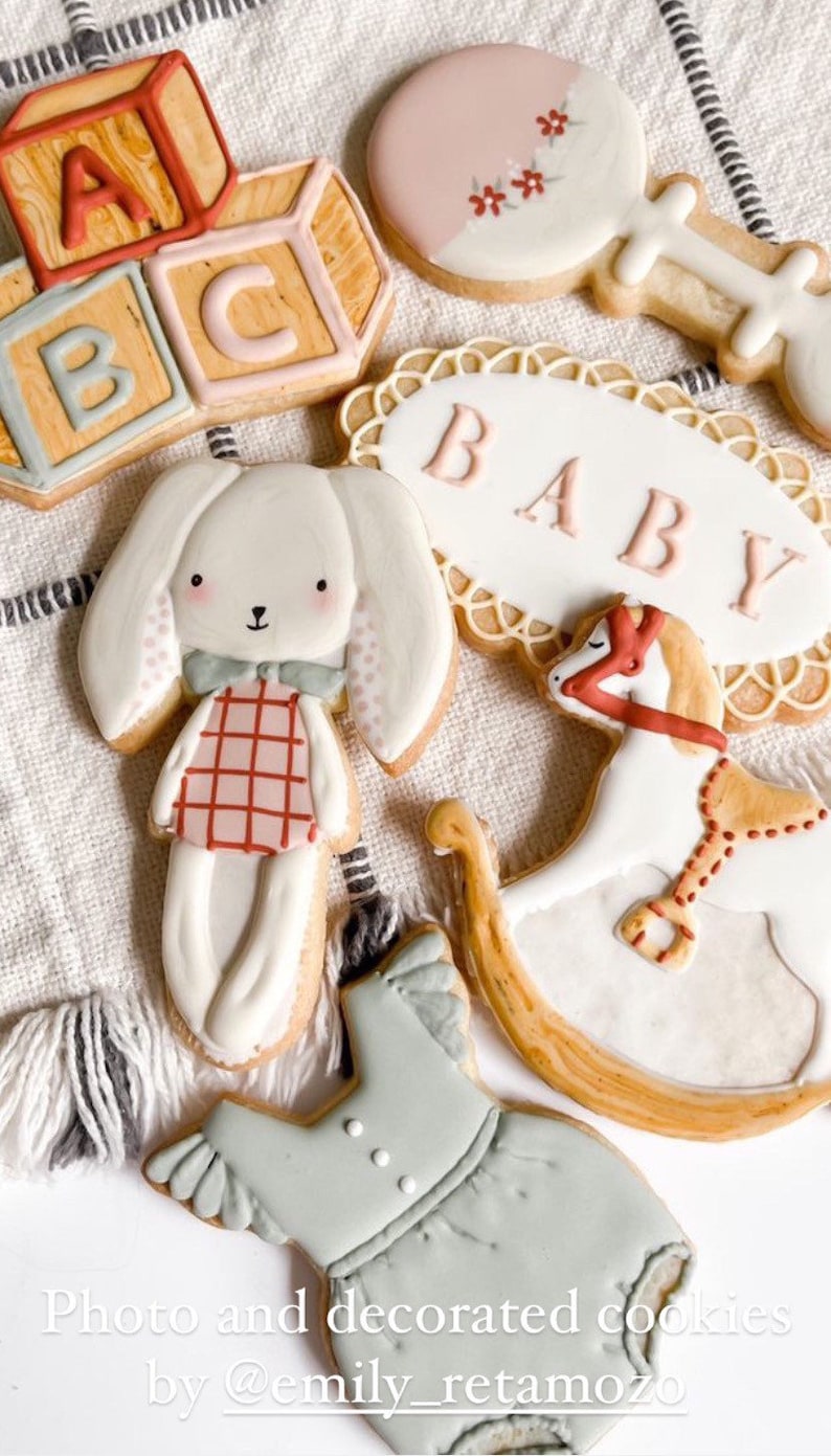 May include: A set of six decorated sugar cookies featuring baby-themed designs. The cookies are decorated with pastel colours and include a rocking horse, a baby rattle, a bunny, a onesie, and blocks that spell "ABC". One cookie has a banner that says "BABY".