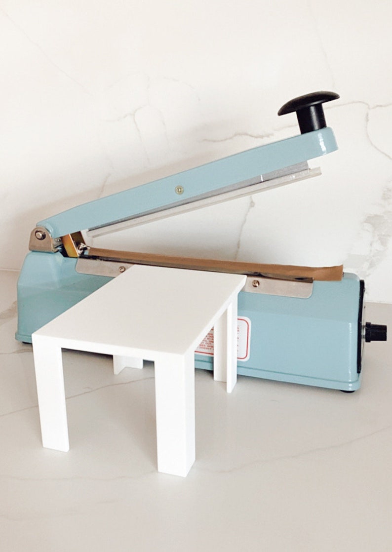 Puede incluir: Un soporte de mesa de pl&aacute;stico blanco con cuatro patas, situado delante de un sellador t&eacute;rmico azul claro con un mango negro. El sellador t&eacute;rmico tiene una tira de papel marr&oacute;n en el interior.