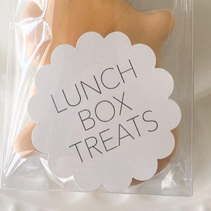 May include: A white scalloped circle sticker with the text "Lunch Box Treats" in black lettering. The sticker is placed on a clear plastic bag with a light brown treat inside.