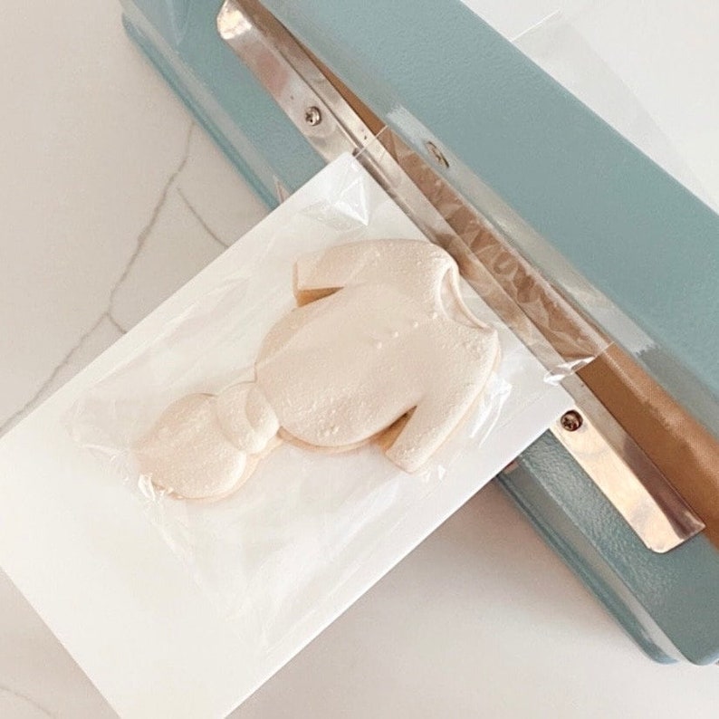 Puede incluir: Una galleta de fondant blanco con forma de body, en una bolsa de pl&aacute;stico transparente, siendo sellada con un sellador t&eacute;rmico azul.