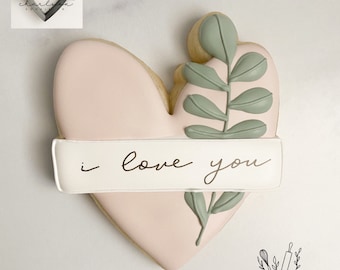 May include: A pink heart-shaped cookie decorated with a green leaf design and a white banner with the text "i love you".