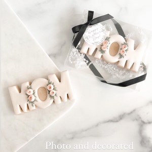 May include: Two decorated sugar cookies spelling "MOM" with pink flowers and green leaves. The cookies are on a white marble surface. The cookie on the left is on a white plate. The cookie on the right is in a clear plastic box with a black ribbon and a round tag that says "Happy Mother's Day!"