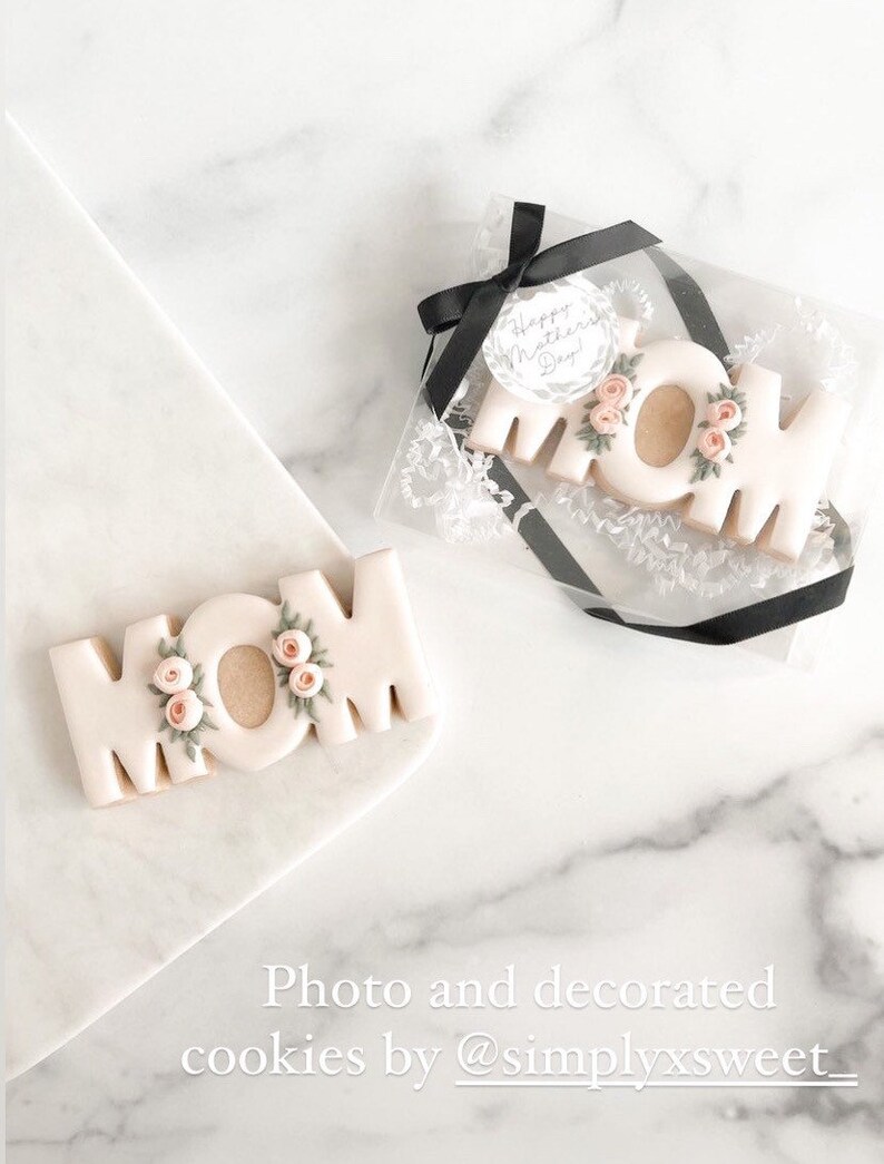 May include: Two decorated sugar cookies spelling "MOM" with pink flowers and green leaves. The cookies are on a white marble surface. The cookie on the left is on a white plate. The cookie on the right is in a clear plastic box with a black ribbon and a round tag that says "Happy Mother's Day!"