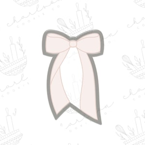 May include: A light pink bow-shaped cookie cutter with a gray outline. The bow has two long, flowing ribbons. The background is white with faint illustrations of baking tools and the text "Charlson Co."
