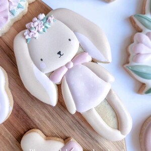 May include: A decorated sugar cookie shaped like a bunny wearing a pink dress and a flower crown. The cookie is on a wooden surface.