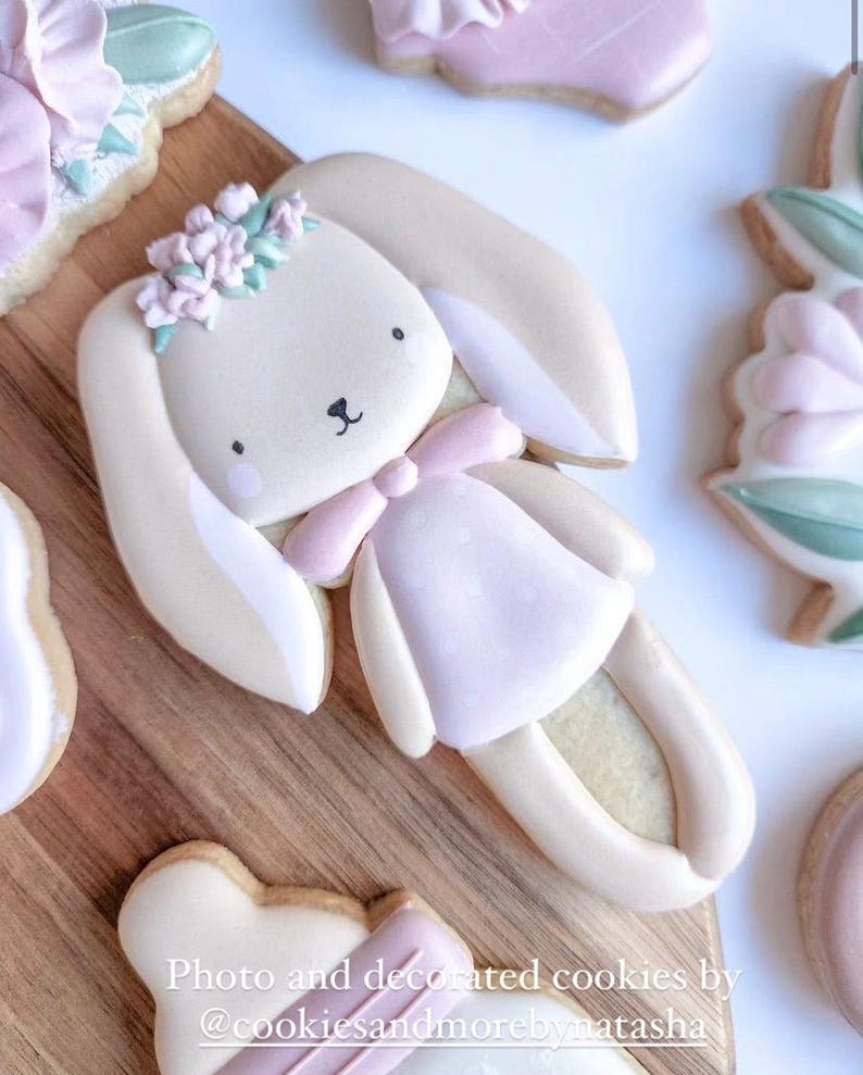 May include: A decorated sugar cookie shaped like a bunny wearing a pink dress and a flower crown. The cookie is on a wooden surface.