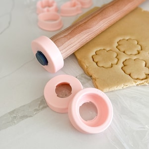 Puede incluir: Rodillo de amasar rosa con mango de madera y cortadores de galletas rosas sobre una superficie blanca. El rodillo tiene una tapa de extremo rosa. Los cortadores de galletas tienen forma de flores y círculos. Hay una hoja de masa para galletas en el fondo.