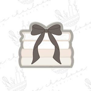 以下が含まれることがあります： 本が積み重なった形にリボンが付いたクッキーカッター。本は白とベージュの色合いで、リボンは濃い茶色です。焼き菓子やテーマのあるクッキーを作るためのカッターです。