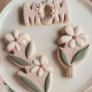 May include: Three decorated sugar cookies on a white plate. One cookie is shaped like the word "MOM" with a floral design. The other two cookies are shaped like flowers with green leaves.