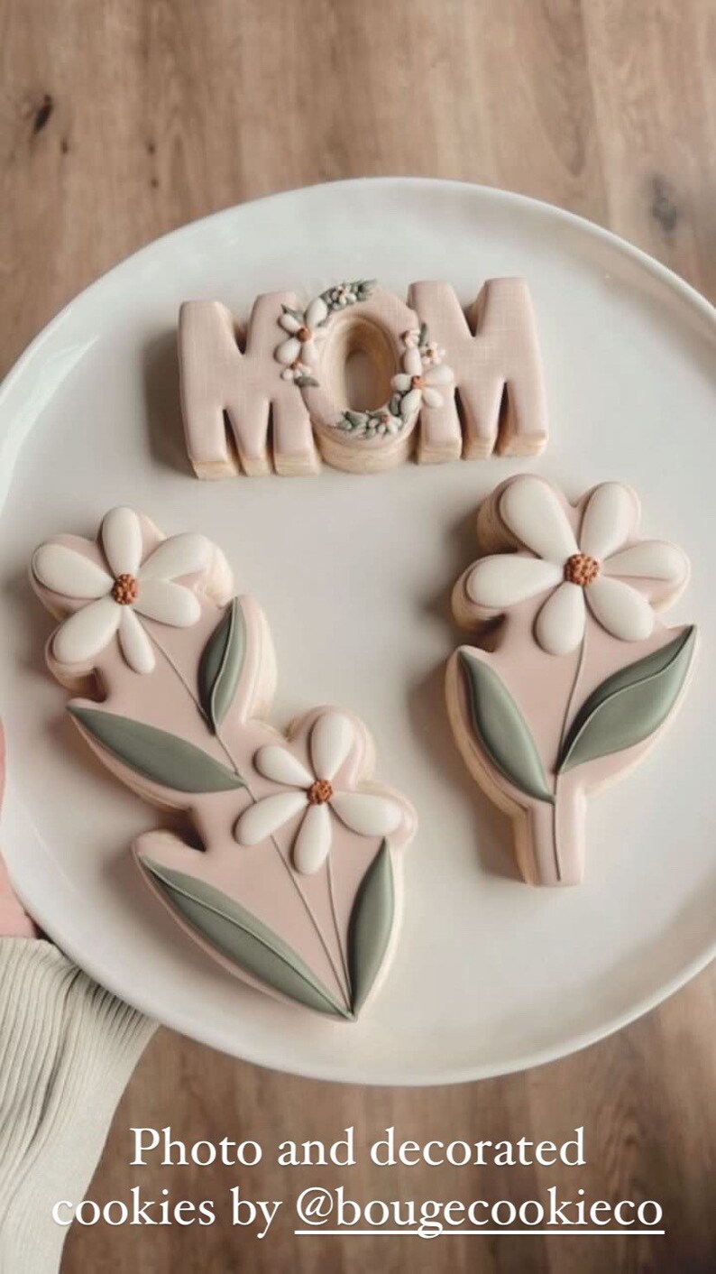 May include: Three decorated sugar cookies on a white plate. One cookie is shaped like the word "MOM" with a floral design. The other two cookies are shaped like flowers with green leaves.