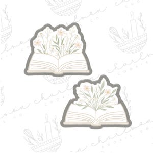 以下が含まれることがあります： 花束が入った開いた本の形をしたクッキーカッターが2つ。カッターは金属製で、グレーの仕上げです。