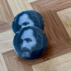 May include: Two black and white button pins featuring a close-up portrait of a man with long hair and a beard.