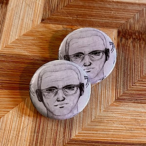 Op de afbeelding: Twee ronde, witte knopen met een zwart-wit illustratie van een man met een bril. De knopen liggen op een houten oppervlak met een visgraatpatroon.