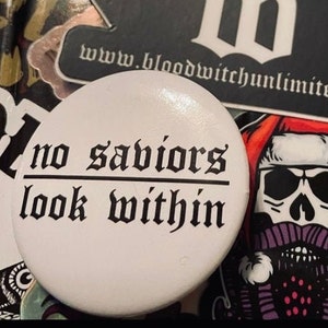 May include: White button with black text that reads "no saviors look within".