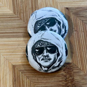 Może przedstawiać: Dwie czarno-białe przypinki z wizerunkiem osoby w bluzie z kapturem i okularach przeciwsłonecznych.