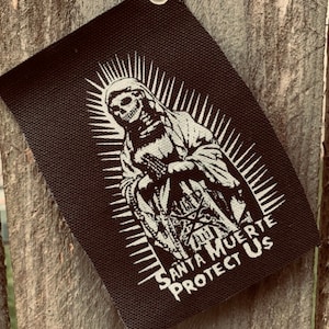 May include: Black and white patch with a skeletal figure of Santa Muerte, a Mexican folk saint, with the text "Santa Muerte Protect Us".