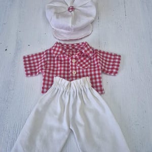 May include: A vintage doll outfit featuring a white cap, a red and white checkered short-sleeved shirt, and white pants. The shirt has three buttons down the front. The outfit is made of fabric and is likely for a small doll.