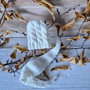 May include: White knitted baby hat and matching scarf with a cable knit pattern. The hat and scarf are lying on a white wooden surface with dried leaves.