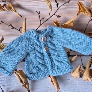 May include: A light blue knitted baby cardigan with a cable knit pattern and wooden buttons.