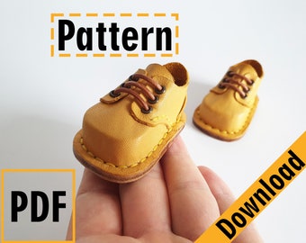 May include: A pair of yellow leather miniature shoes with brown laces and stitching. The shoes are on a hand and the text "Pattern: PDF Download" is in the background.