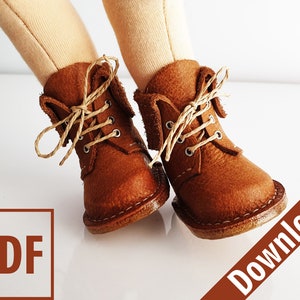 Puede incluir: Primer plano de un par de pequeñas botas de cuero marrón con cordones color beige. Las botas tienen un diseño clásico con costuras y ojales visibles. Las botas están sobre una superficie blanca. También contiene el texto "PDF" y "Download".