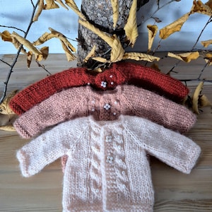May include: Three hand-knitted baby sweaters in shades of red, pink, and light pink. Each sweater features a cable knit design on the front and is adorned with small, white pearl-like buttons. The sweaters are arranged on a wooden surface.