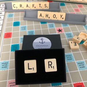 May include: A close-up of a black box containing two Scrabble tiles. The tiles are cream-coloured with black lettering. The left tile has the letter "L" and the number "1" on it. The right tile has the letter "R" and the number "1" on it. The box is sitting on a Scrabble game board.