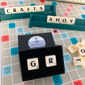 May include: A black box containing two white Scrabble tiles, one with the letter "G" and the number "2" and the other with the letter "R" and the number "1". The box is on a beige and pink game board with the word "CRAFTS" and "AHOY" spelled out in white tiles.