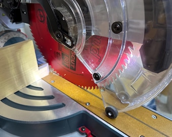 May include: A red circular saw blade with 80 teeth is mounted on a miter saw, partially covered by a clear plastic safety guard. A light-colored wooden board is positioned against the metal fence, ready for a cut on the gray metal base.