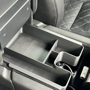 May include: A black plastic car centre console organiser with multiple compartments. The organiser is installed in a car and has a black cord running through it.