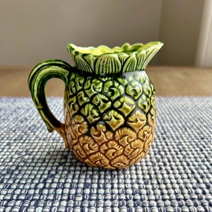 May include: A ceramic pitcher shaped like a pineapple with green and brown glaze. The pitcher has a handle and a spout.