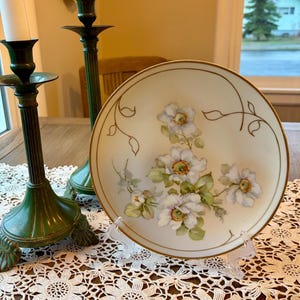 May include: A decorative plate with a cream-colored background and a gold rim. The plate features a floral design with white flowers, green leaves, and brown stems. Two green candle holders with white candles are in the background.
