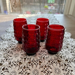 May include: Four ruby red drinking glasses with a textured, bubble-like pattern. The glasses are cylindrical with a slightly flared rim and sit on a decorative, openwork lace doily. The glasses are empty and appear to be made of glass.