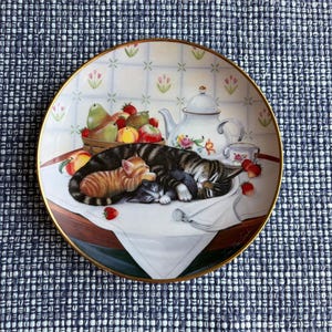 May include: A round decorative plate with a gold rim featuring an illustration of two sleeping cats, one orange tabby and one grey, resting on a white cloth. The scene includes a basket of fruit, a white teapot, and a small cup on a table.