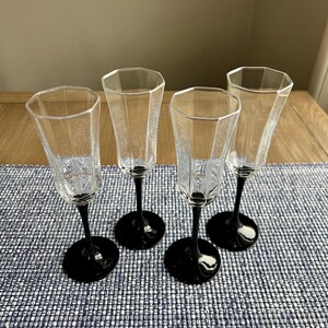 May include: Four clear glass champagne flutes with black stems. The flutes have a geometric, faceted design.