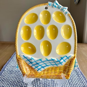 May include: A white ceramic egg plate shaped like a basket with a blue and white checkered tablecloth. The plate has eight yellow egg-shaped indentations.