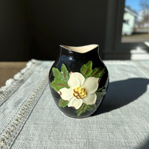 Puede incluir: Una pequeña jarra de cerámica negra con un diseño de flor blanca. La flor tiene hojas verdes y un centro amarillo.