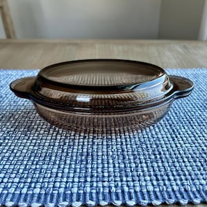 May include: A brown glass casserole dish with a matching lid. The dish has two handles and is sitting on a blue and white woven placemat. The dish is made of a transparent material.