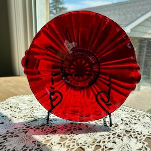 May include: A red glass plate with a ruffled edge and a raised center design. The plate is on a black metal stand.