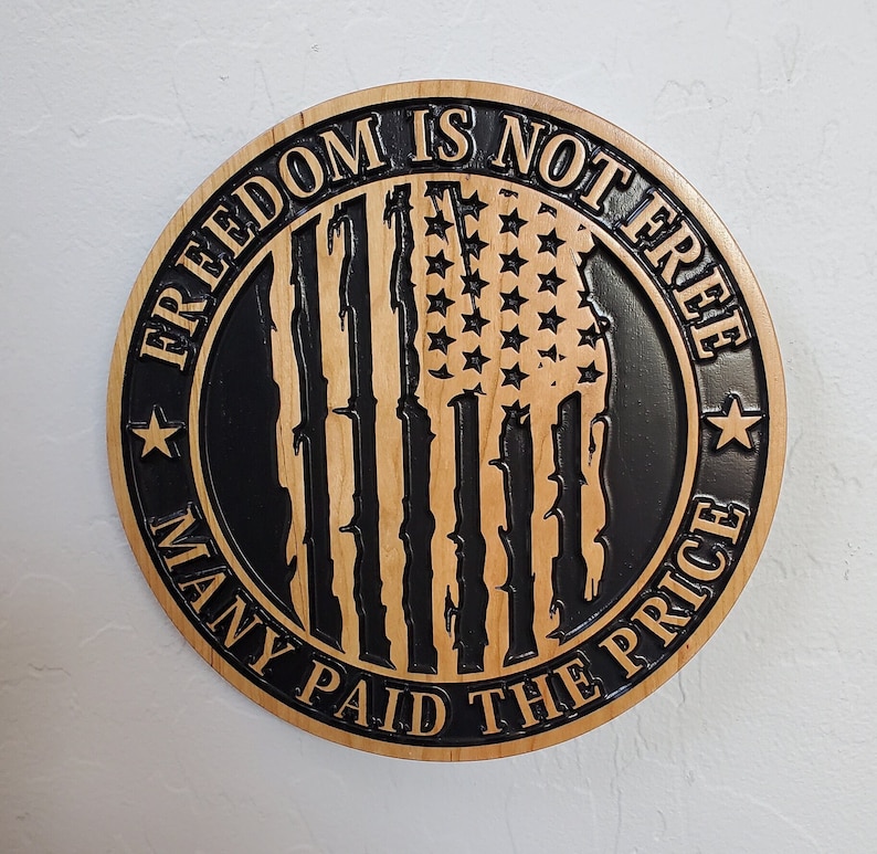 K&ouml;nnte beinhalten: Ein rundes Holzschild mit dem Text "Freedom is not free many paid the price" in einem schwarzen Kreis mit einem verwitterten amerikanischen Flaggenmuster in der Mitte.