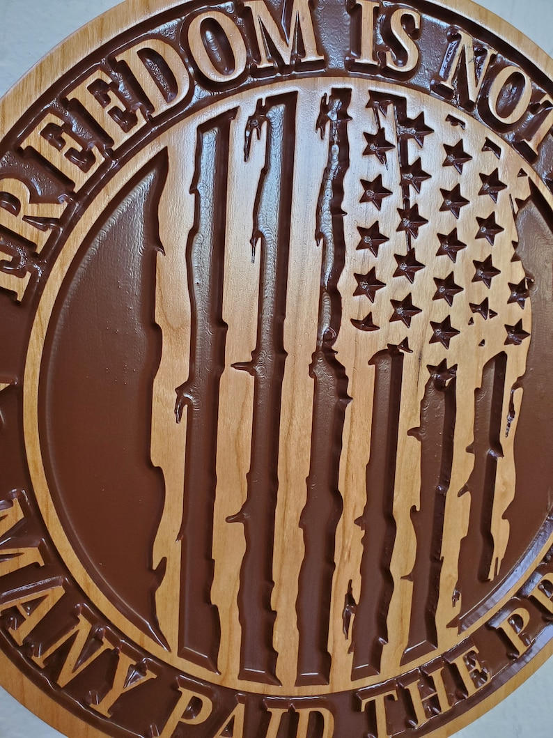 K&ouml;nnte beinhalten: Ein rundes Holzschild mit einer geschnitzten amerikanischen Flagge und dem Text "Freiheit ist nicht umsonst, viele haben den Preis bezahlt".