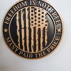 K&ouml;nnte beinhalten: Ein rundes Holzschild mit einem schwarz-wei&szlig;en amerikanischen Flaggen-Design. Der Text "Freedom is not free many paid the price" ist um die Flagge herum geschrieben.