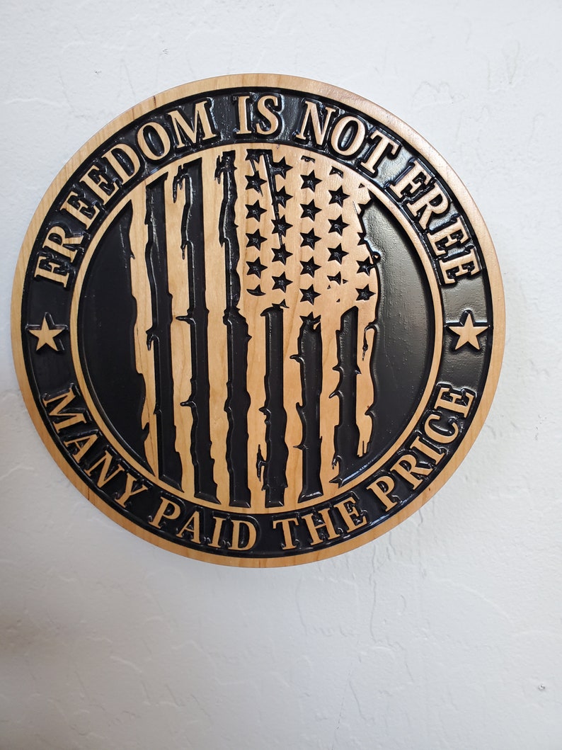 K&ouml;nnte beinhalten: Ein rundes Holzschild mit einem schwarz-wei&szlig;en amerikanischen Flaggen-Design. Der Text "Freedom is not free many paid the price" ist um die Flagge herum geschrieben.