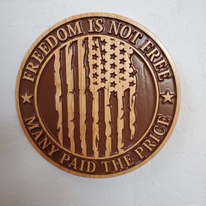 K&ouml;nnte beinhalten: Ein rundes Holzschild mit braunem Hintergrund und einer geschnitzten amerikanischen Flagge in der Mitte. Das Schild lautet "Freiheit ist nicht umsonst, viele haben den Preis bezahlt".