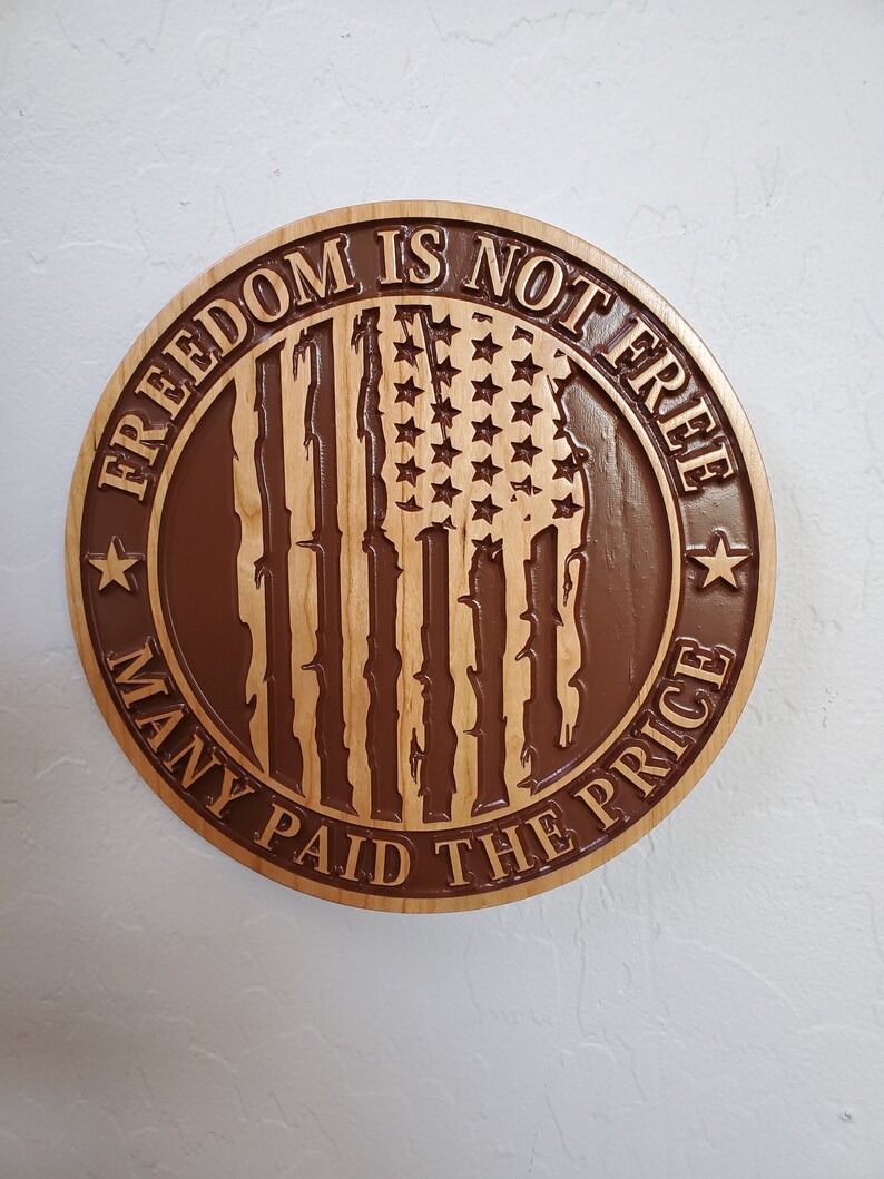 K&ouml;nnte beinhalten: Ein rundes Holzschild mit braunem Hintergrund und einer geschnitzten amerikanischen Flagge in der Mitte. Das Schild lautet "Freiheit ist nicht umsonst, viele haben den Preis bezahlt".
