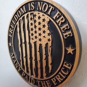 K&ouml;nnte beinhalten: Ein rundes Holzschild mit schwarzem Hintergrund und einem verwitterten amerikanischen Flaggenmuster. Der Text "Freedom is not free, many paid the price" ist in das Holz geschnitzt.